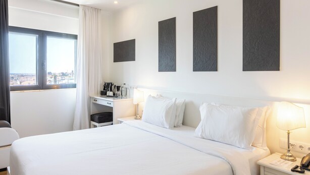 Habitación de hotel con una cama grande, un escritorio con cafetera, una lámpara y vistas a la ciudad a través de una ventana.