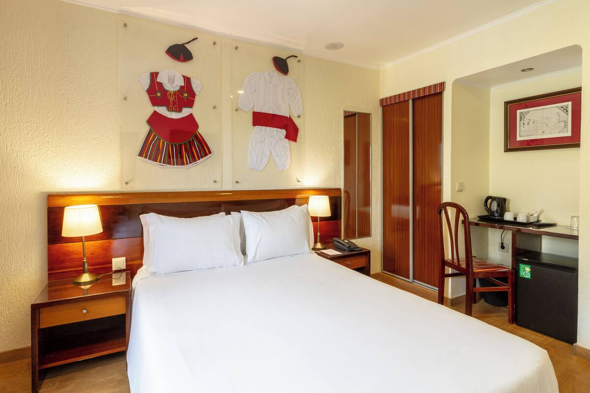 A hotel room with a large bed, two bedside tables, a desk with a chair, and a coat rack with clothes.