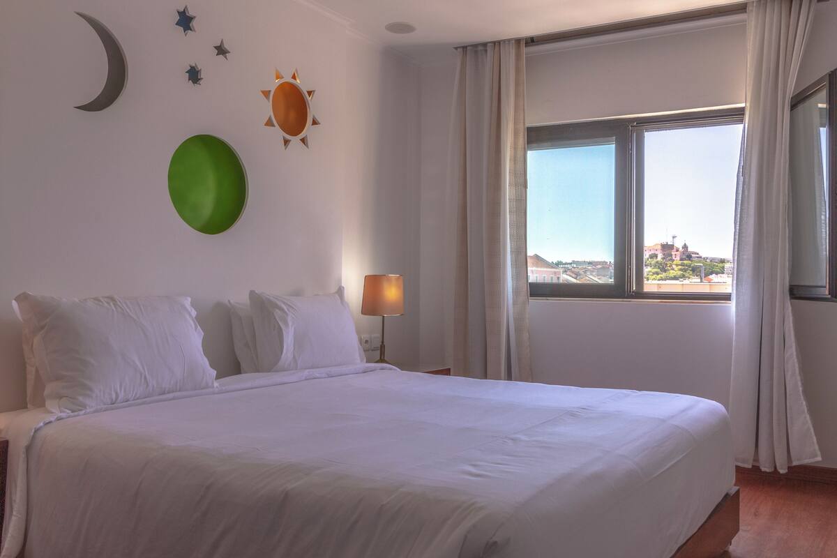 A hotel room with a large bed, a nightstand with a lamp, a window with curtains, and wall decorations including a crescent moon, stars, and a sun.