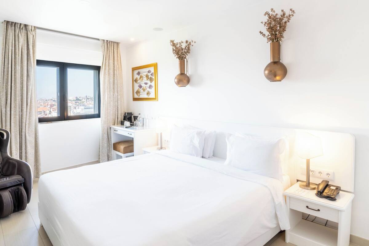A hotel room with a bed, a massage chair, a desk, a nightstand with a lamp, and a view of the city through the window.