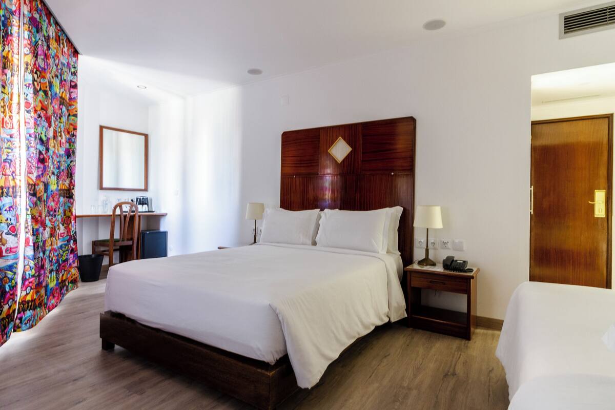 A hotel room with a large bed, wooden headboard, bedside tables with lamps, a desk with a chair, and a colorful curtain.