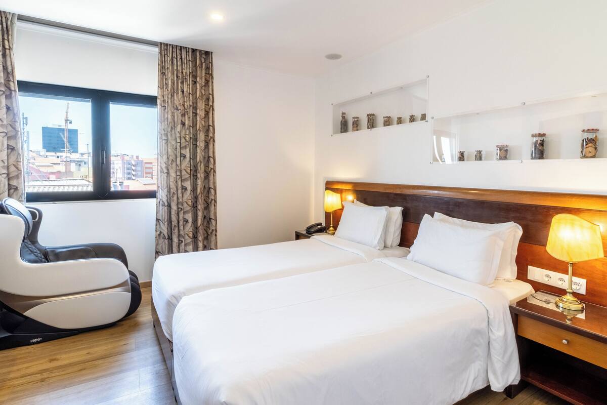 A hotel room with two beds, a massage chair, a bedside table with a lamp, and a city view through a large window.