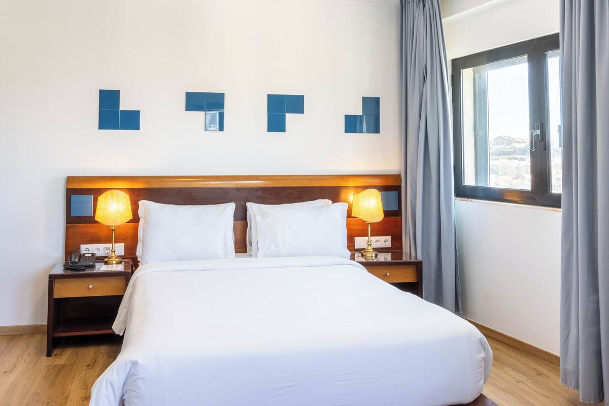 A hotel room with a large bed, two bedside tables with lamps, a wooden headboard, and a window with curtains.