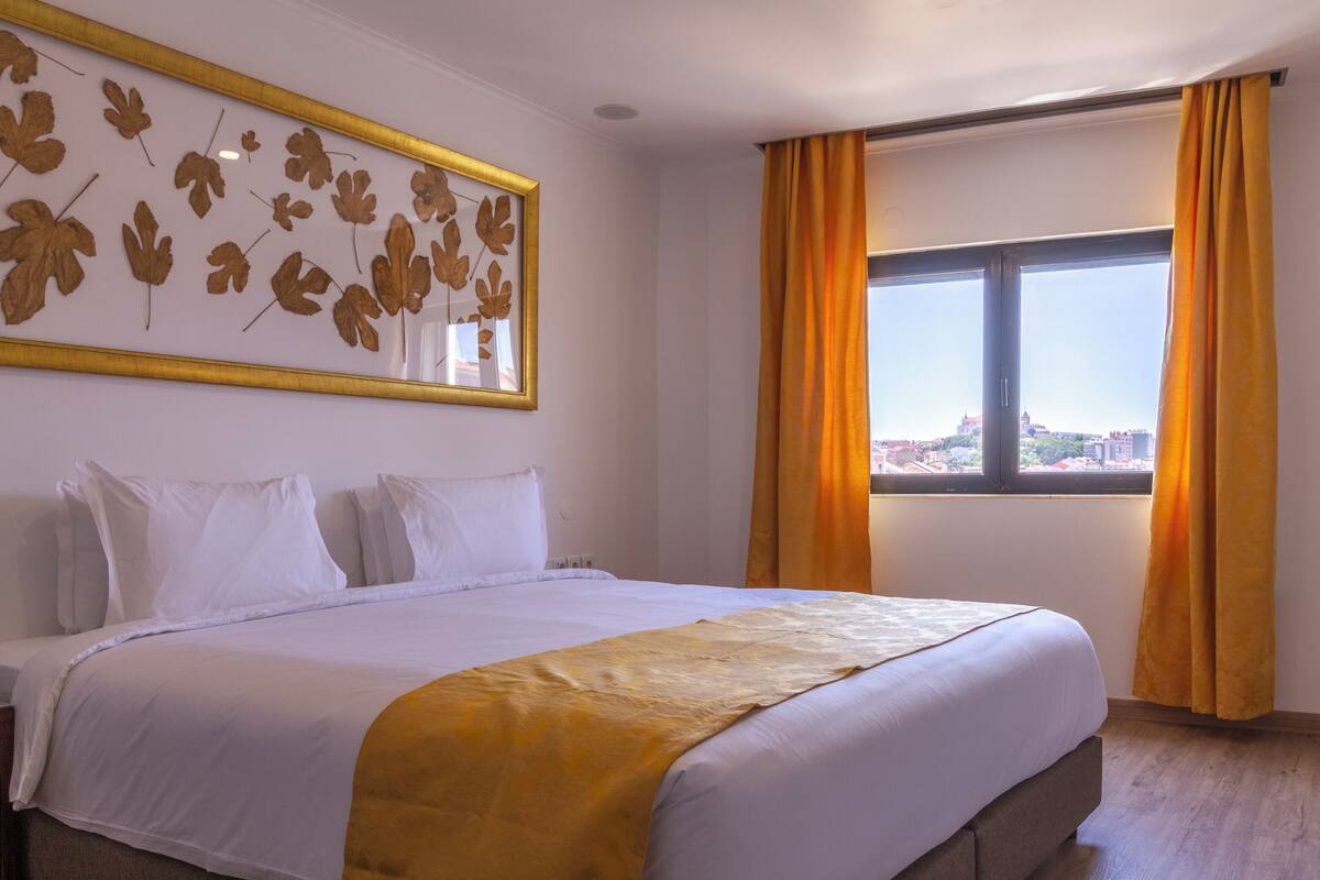 A hotel room with a large bed, a view of the city through a window, and a wall-mounted leaf decoration.