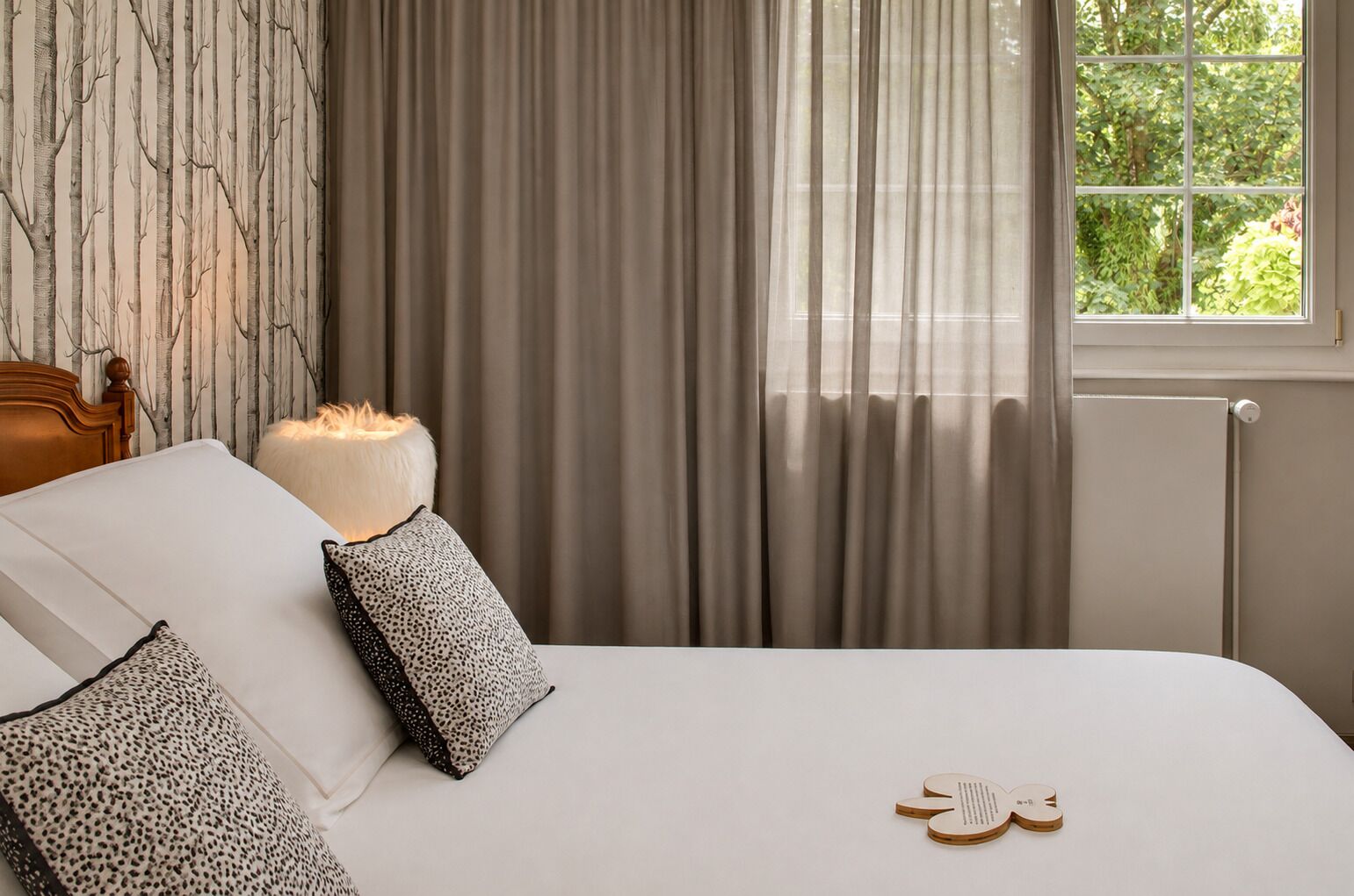 Une chambre à coucher avec un lit, des oreillers, une table de chevet, une lampe et une fenêtre avec des rideaux.