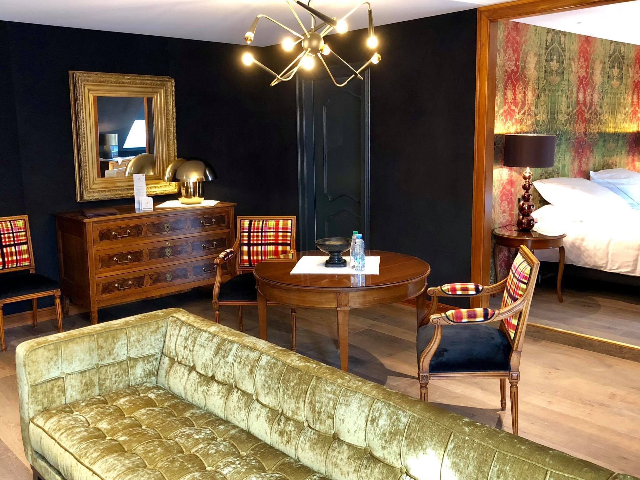 A hotel room with a round wooden table, a sofa, two chairs, a dresser, a bed, and a chandelier.