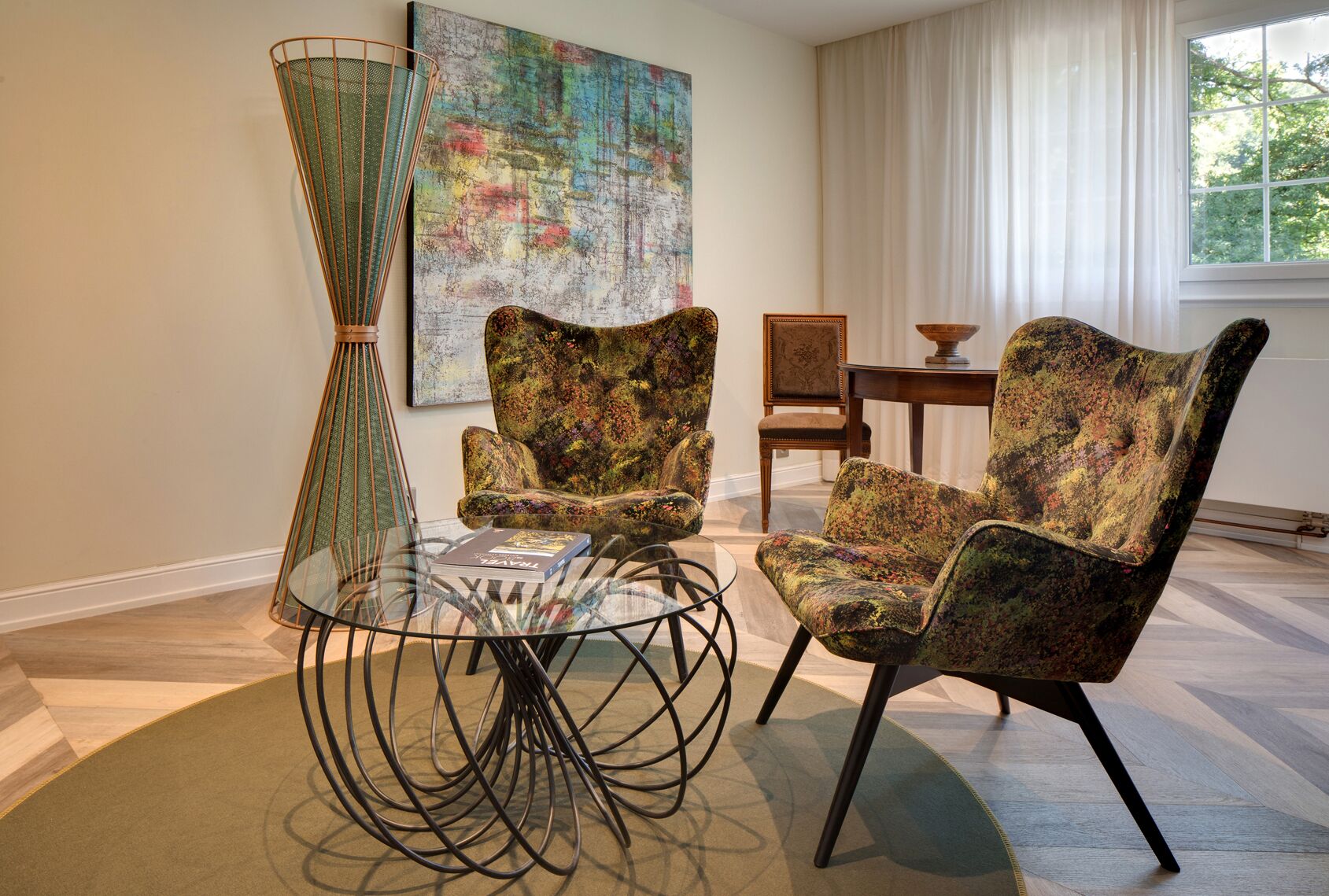 A modern living room with a glass coffee table, two patterned armchairs, a decorative floor lamp, and a large abstract painting on the wall.
