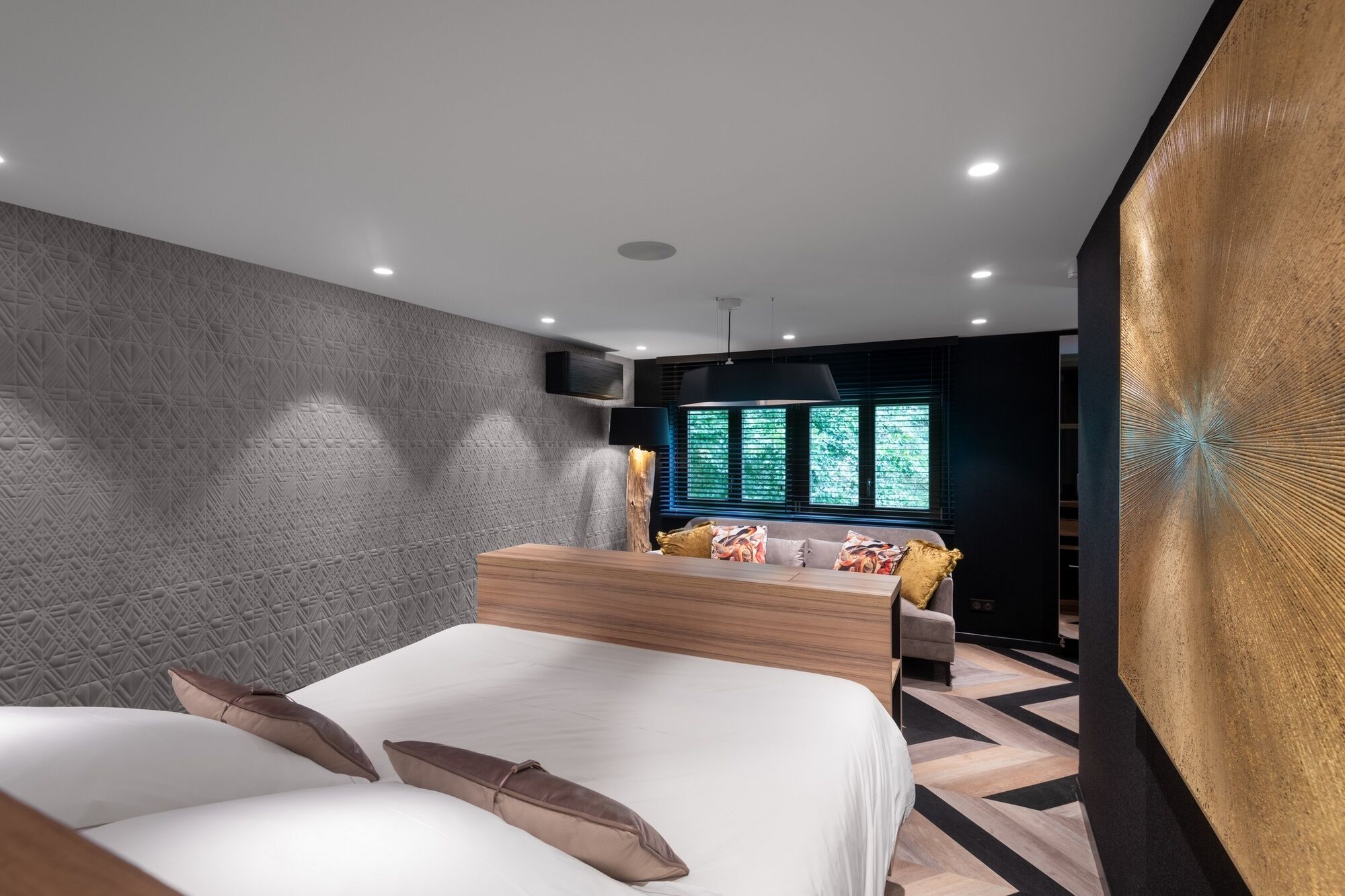 Un dormitorio moderno con una cama grande, una zona de estar con sofá y sillones, un televisor montado en la pared y una ventana con persianas.