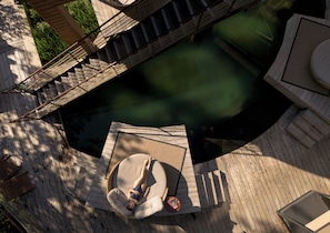 Una persona recostada en una silla hamaca junto a una piscina, rodeada por una terraza de madera y un puente.