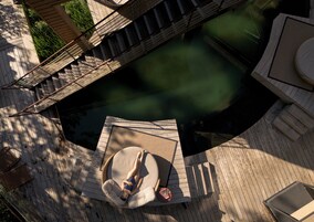 Una persona recostada en una silla hamaca junto a una piscina, rodeada por una terraza de madera y un puente.