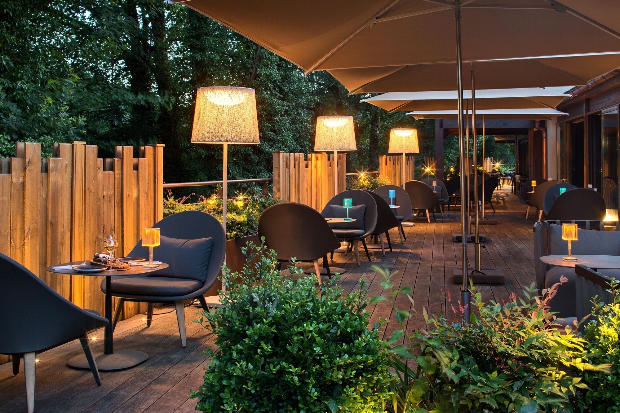 An outdoor patio with wooden furniture and tables, surrounded by greenery and illuminated by lamps.