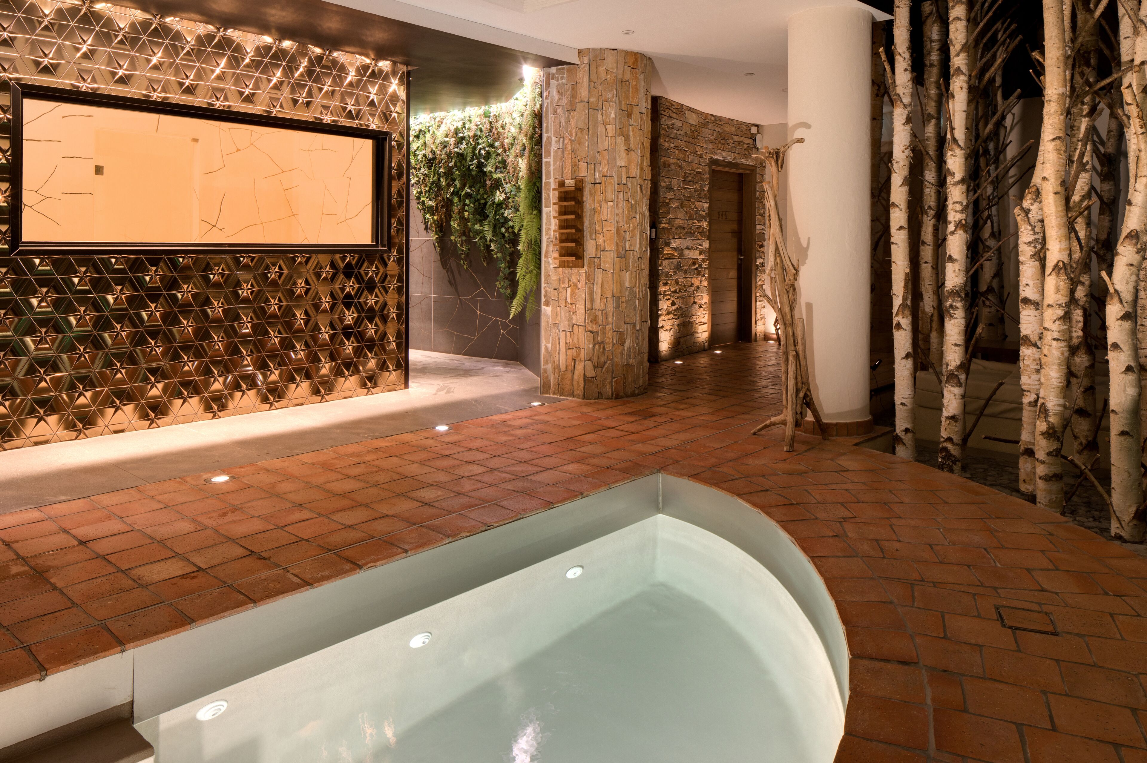 A spa area with a glass-enclosed hot tub, a wall with a geometric pattern, and a stone wall with greenery.
