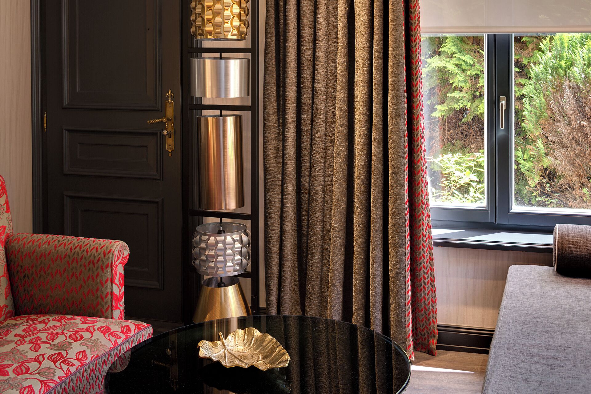A room with a dark wooden door, a red patterned sofa, a black table with a gold decorative item, and a window with curtains.