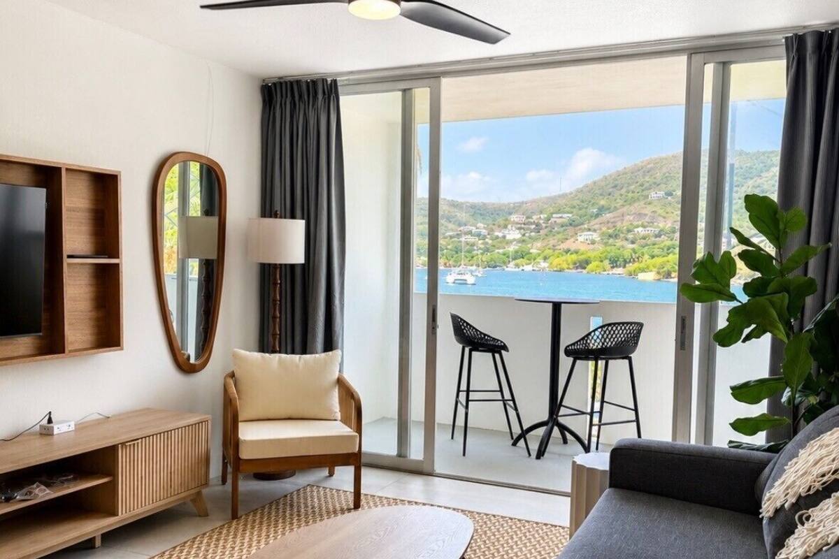 A modern hotel room with a sofa, a TV, a ceiling fan, a balcony with chairs, and a view of the sea.