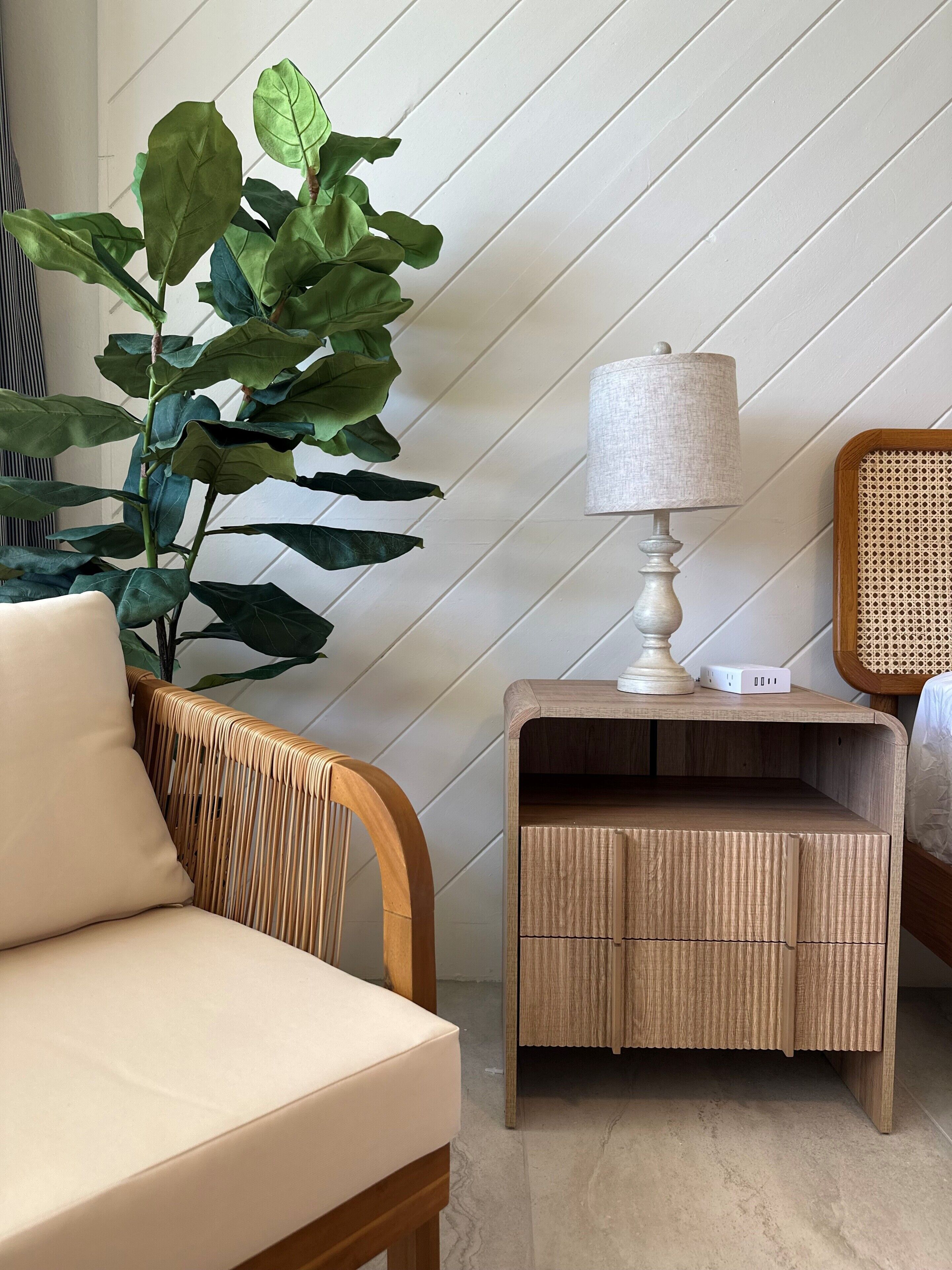A room with a wicker chair, a bedside table with a lamp, and a large plant.