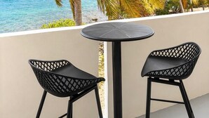 Un balcón con una mesa redonda y dos sillas, con vista a una playa con palmeras y un mar azul transparente.