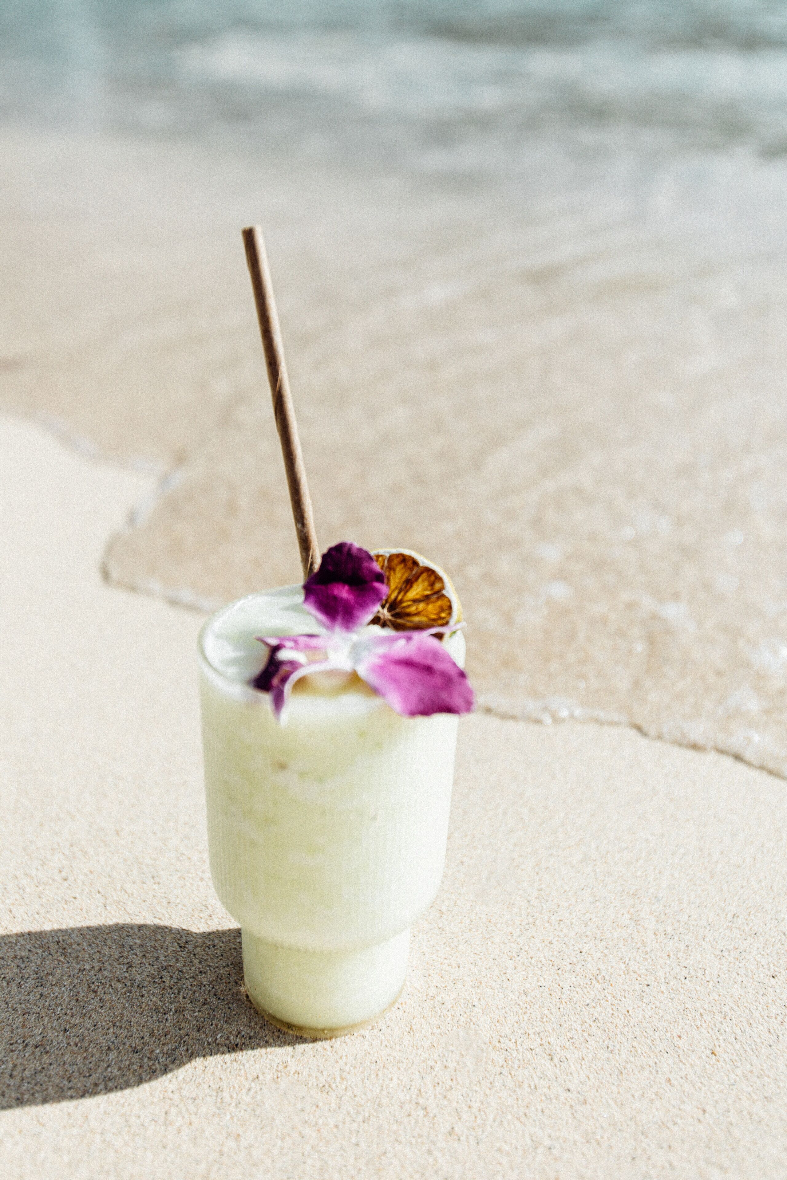 Un verre d’une boisson crémeuse, agrémenté d’une paille et d’une fleur violette, posé sur une plage de sable.