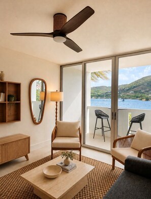 Una habitación con ventilador de techo, un espejo grande, una consola de madera, un sofá, una mesa de centro y vista a una masa de agua a través de puertas correderas amplias.