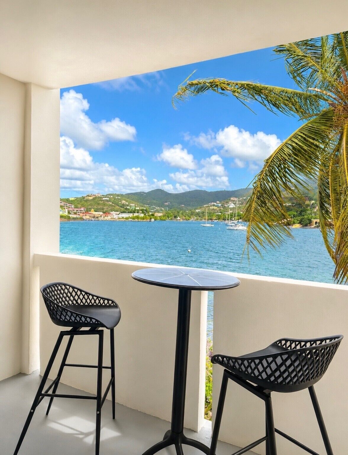 Ein Balkon mit zwei schwarzen Stühlen und einem kleinen runden Tisch, mit Blick auf ein Gewässer mit Booten und einer Stadt in der Ferne.
