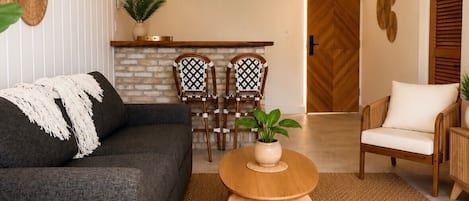Una sala moderna con un sofá, una mesa de centro redonda de madera, un reposapiés blanco, un área de comedor con sillas, un ventilador de techo y una zona de cocina.