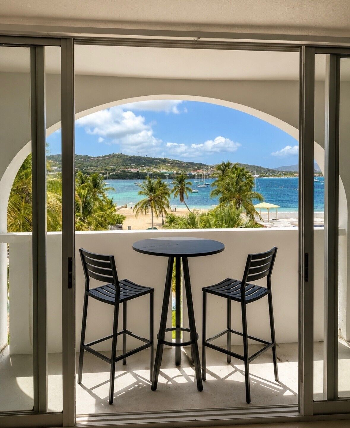 Ein Balkon mit Tisch und zwei Stühlen, mit Blick auf einen Strand mit Palmen und einem klaren Himmel.
