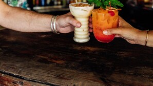 Un bar con estantes de madera llenos de botellas, una barra de madera y dos manos sosteniendo bebidas.