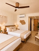 A hotel room with two beds, a ceiling fan, an air conditioning unit, macrame wall decor, and a wooden door.