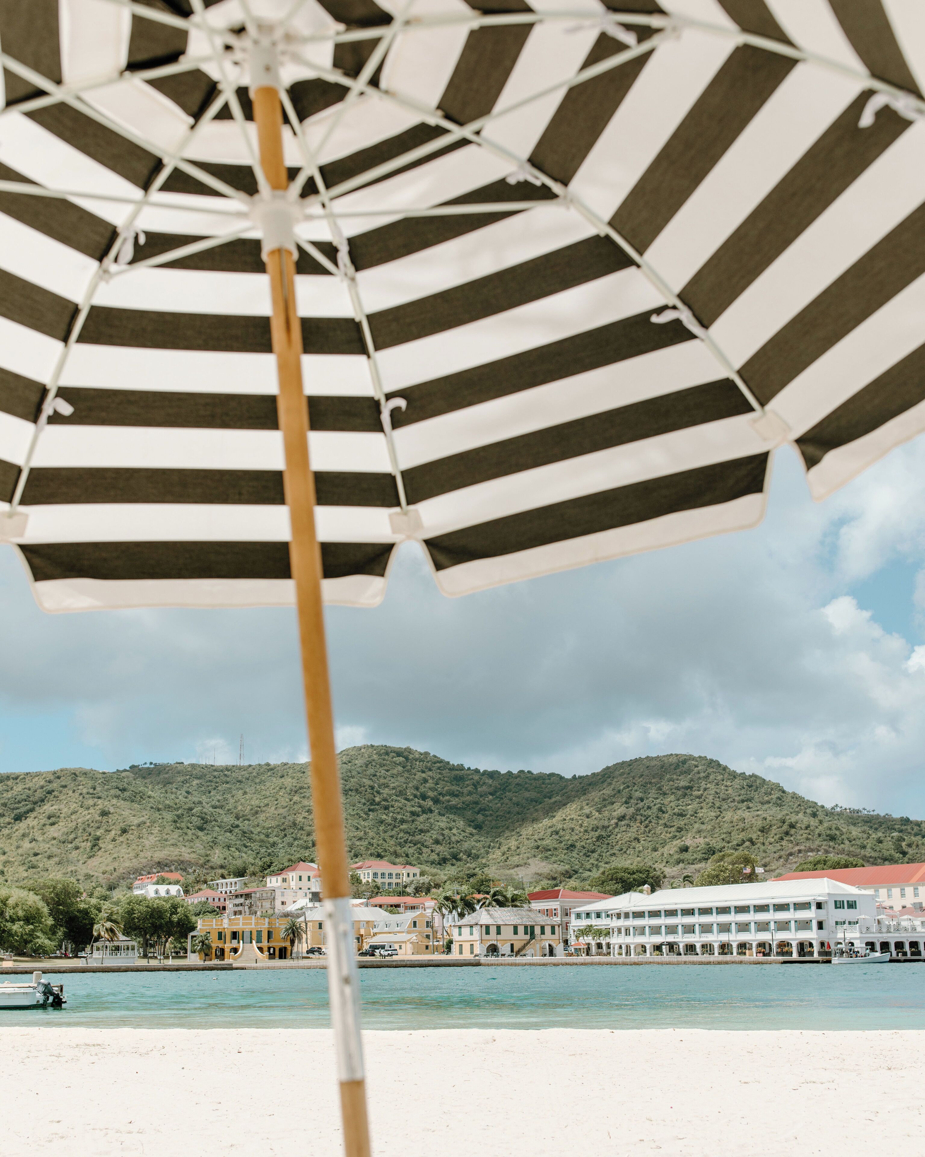 A beach scene with a striped umbrella, a boat, and buildings in the background.