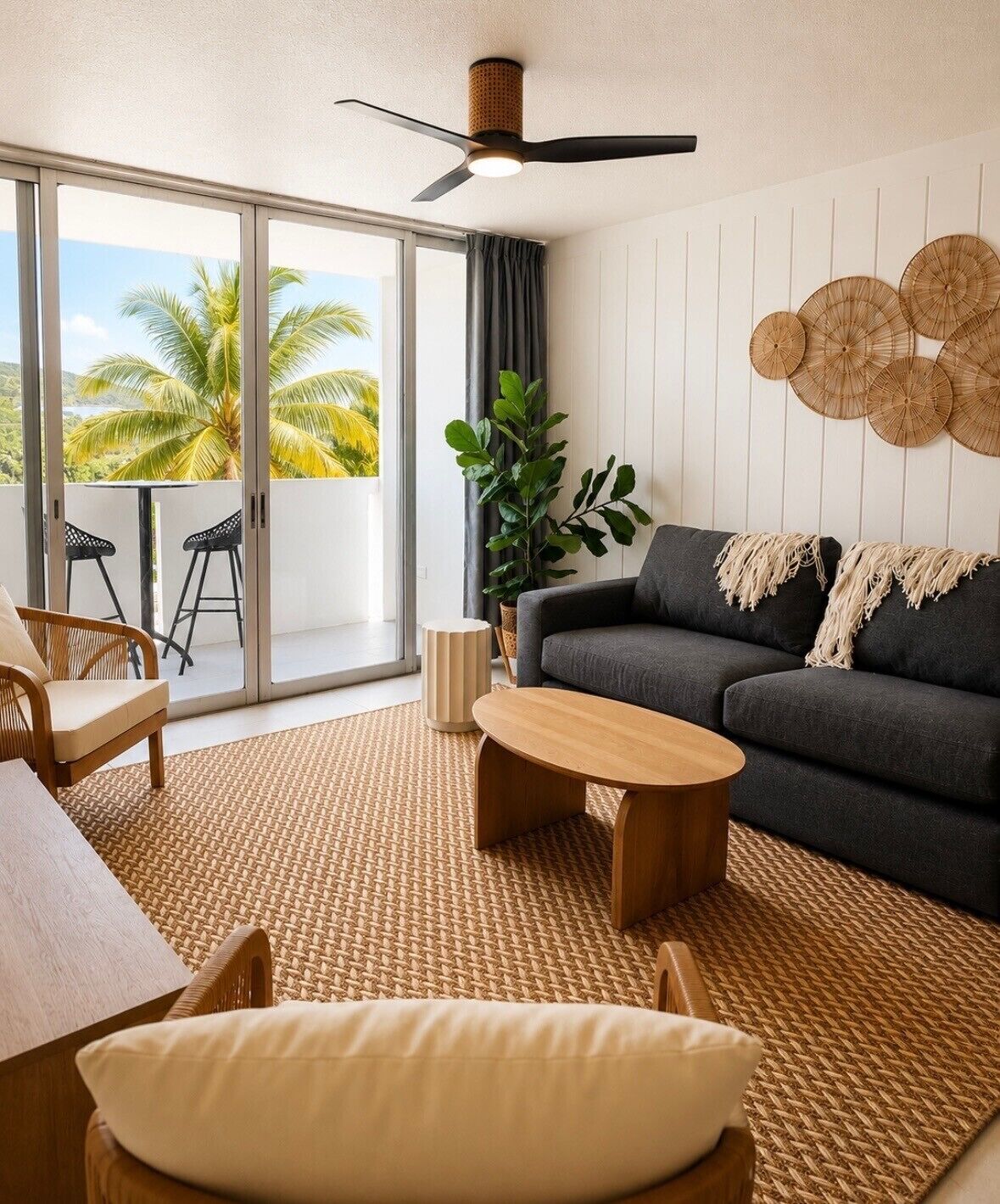 Una sala de estar moderna con sofá, mesa de centro y sillones, un ventanal con vistas a palmeras y un ventilador de techo.