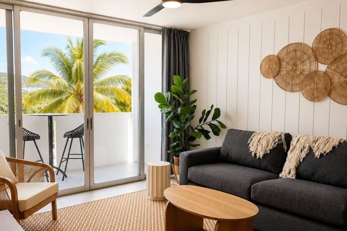 A modern living room with a sofa, coffee table, and chairs, a large window with a view of palm trees, and a ceiling fan.