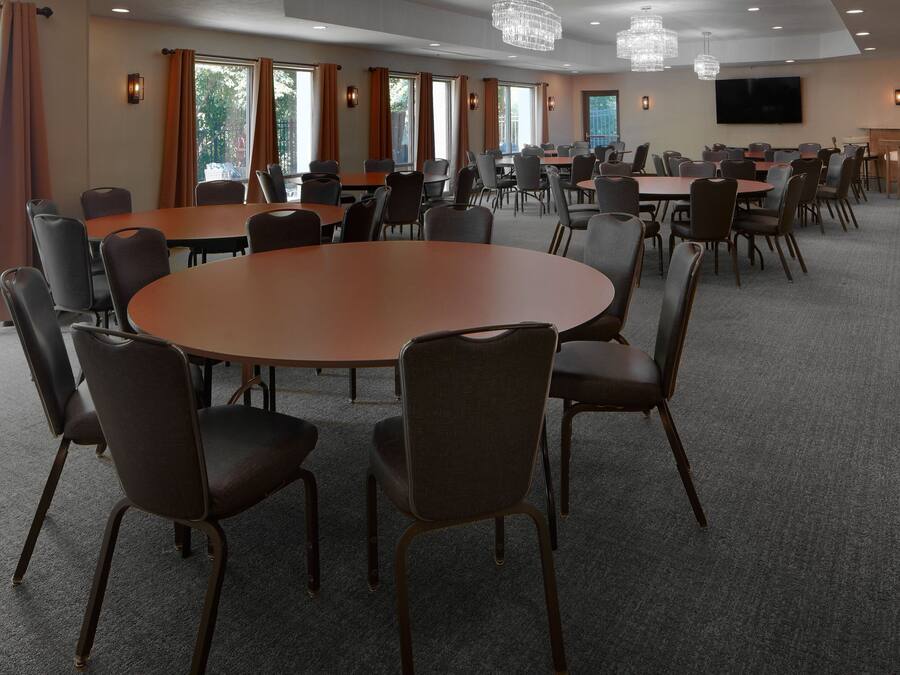 Un salón de conferencias con mesas y sillas redondas, ventanas amplias con cortinas y un televisor de pantalla plana en la pared.