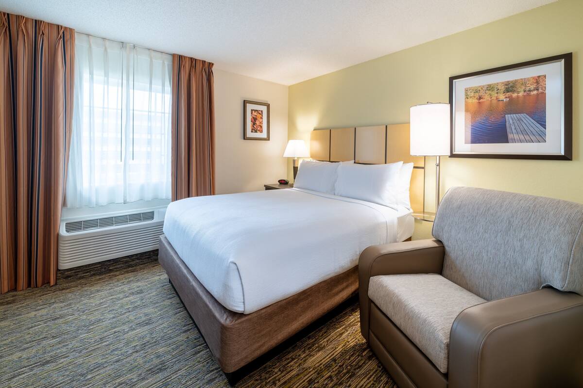 Une chambre d’hôtel avec un grand lit, un canapé, une fenêtre avec des rideaux, une lampe fixée au mur et un tableau encadré.