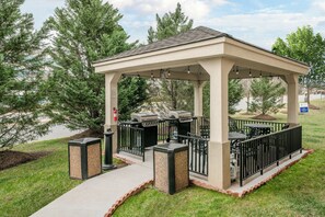 Un espace extérieur abrité, doté d’un barbecue et de sièges, entouré d’arbres et agrémenté d’un panneau indiquant un sentier pédestre.