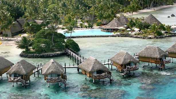 Overwater bungalows with thatched roofs, clear turquoise water, and a palm tree-lined beach.