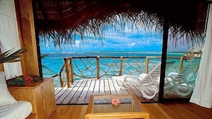 A wooden deck with a view of the ocean, white lounge chairs, and a small table with a flower.