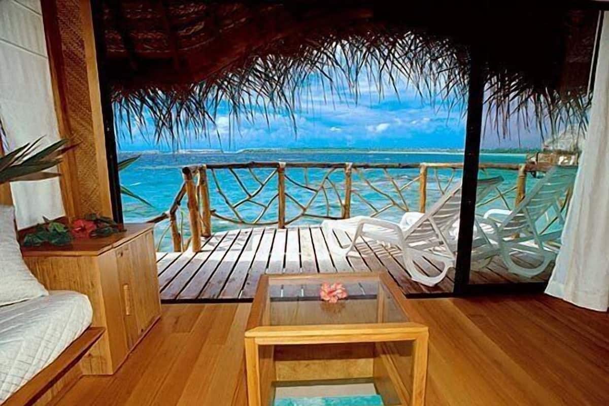 A wooden deck with a view of the ocean, white lounge chairs, and a small table with a flower.