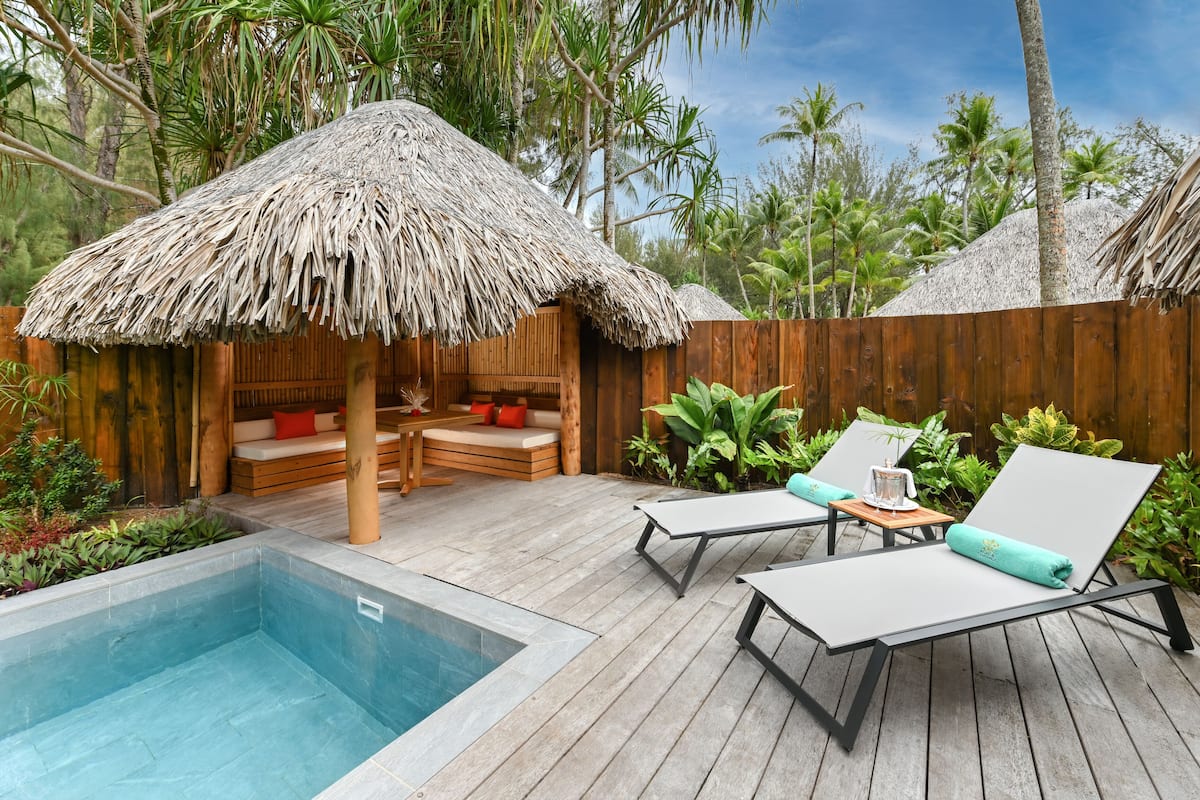 Un terrazzo in legno con piscina, sedie a sdraio, una capanna con tetto di paglia e piante tropicali.