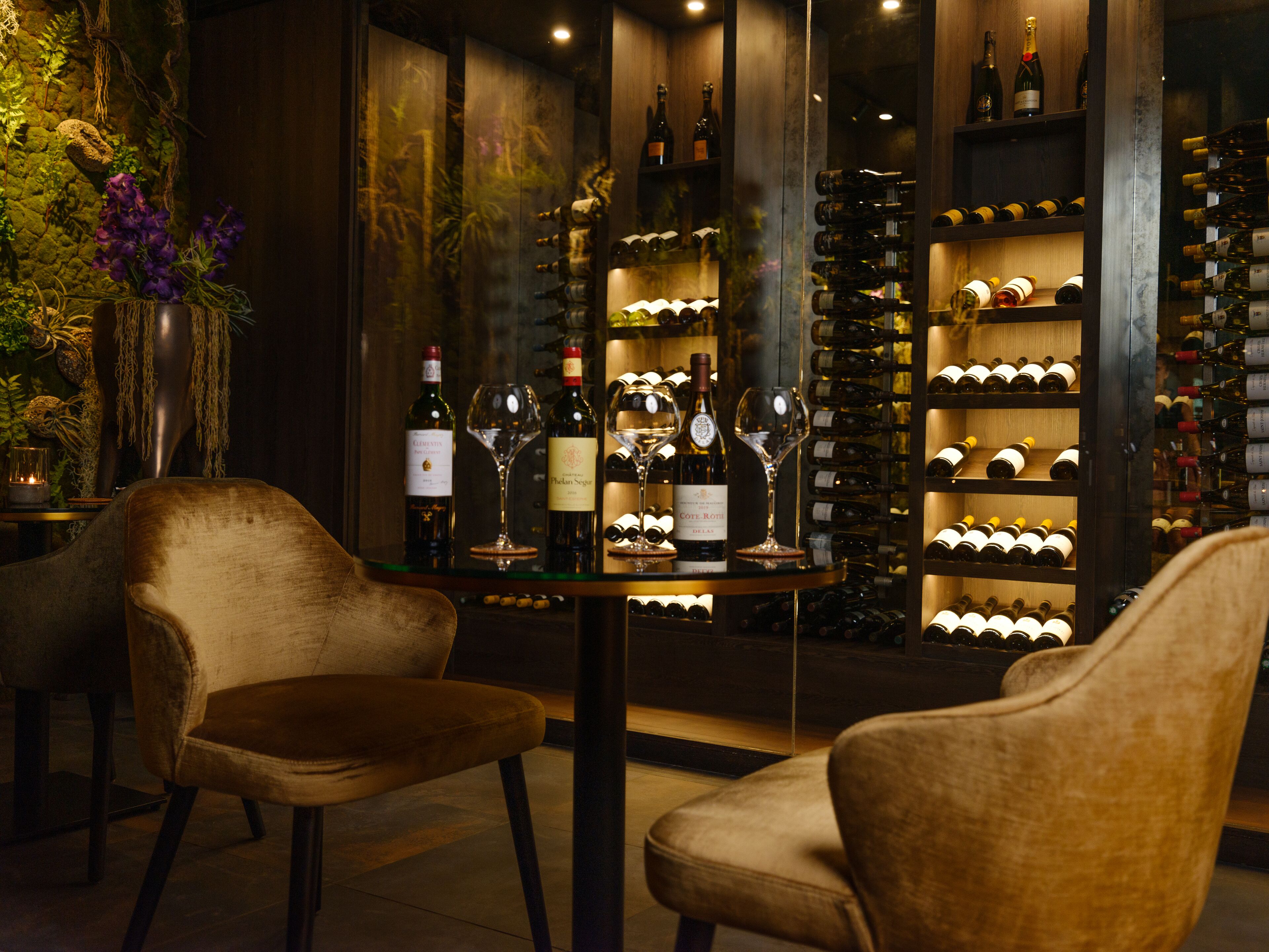 A wine cellar with a selection of wine bottles and glasses on shelves, two velvet chairs, and a decorative plant.