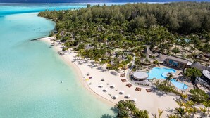 An aerial view of a tropical resort with a beach, clear turquoise waters, and a swimming pool surrounded by palm trees and lounge areas.