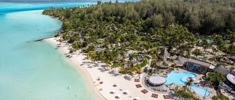 An aerial view of a tropical resort with a beach, clear turquoise waters, and a swimming pool surrounded by palm trees and lounge areas.