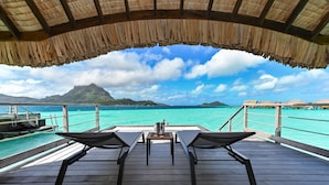 An overwater bungalows with thatched roofs, wooden deck, and lounge chairs facing a turquoise sea and a mountain in the distance.