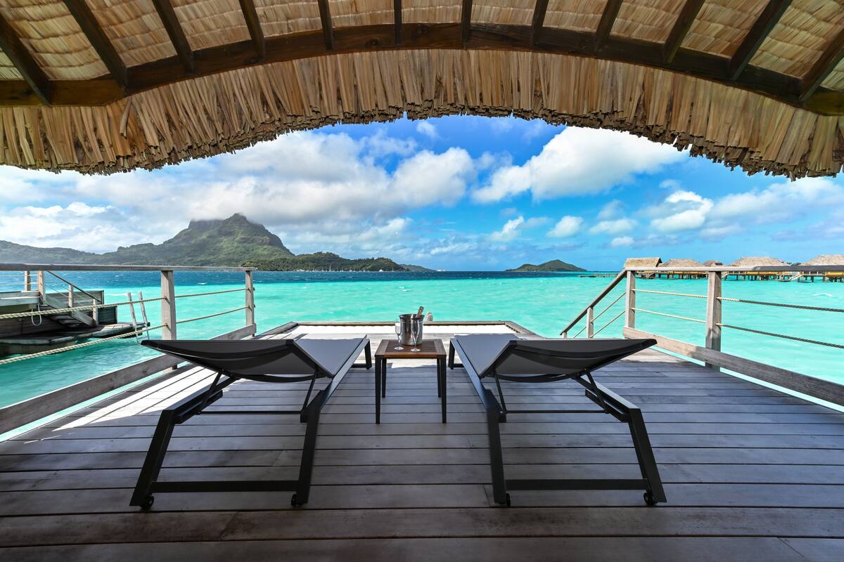 Bungalow sull'acqua con tetti in paglia, veranda in legno e poltrone relax che si affacciano su un mare turchese e una montagna all'orizzonte.