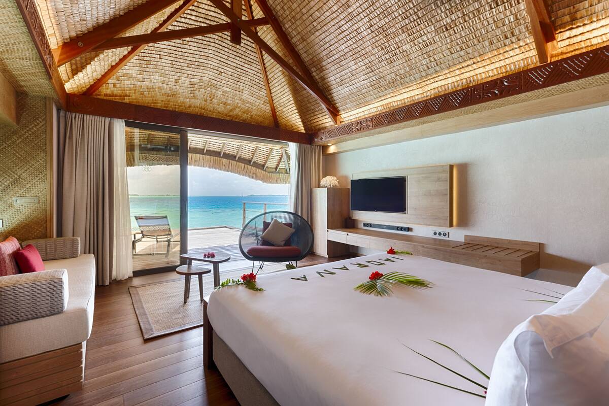 Camera d'albergo con un letto grande, una TV, un'area salotto vista oceano e un balcone con tetto di paglia.