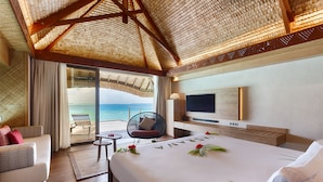A hotel room with a large bed, a TV, a seating area with a view of the ocean, and a balcony with a thatched roof.