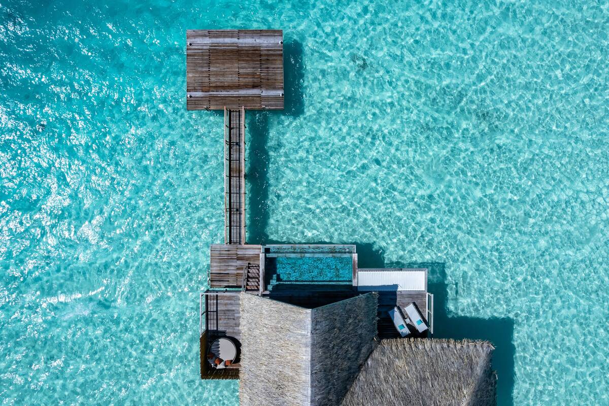 Vista aerea di un resort con una struttura sull'acqua in legno, una piscina e una zona con tetto di paglia.
