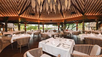 A restaurant with round tables, wicker chairs, and a thatched roof.