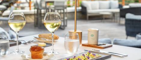 Un espacio exterior para comer con una mesa dispuesta para dos personas, que incluye un postre, dos copas de vino blanco, un muffin y una vela.