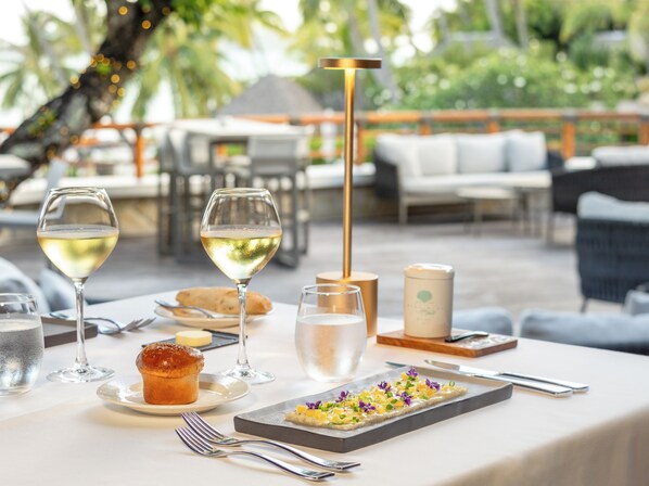 An outdoor dining area with a table set for two, featuring a dessert, two glasses of white wine, a muffin, and a candle.