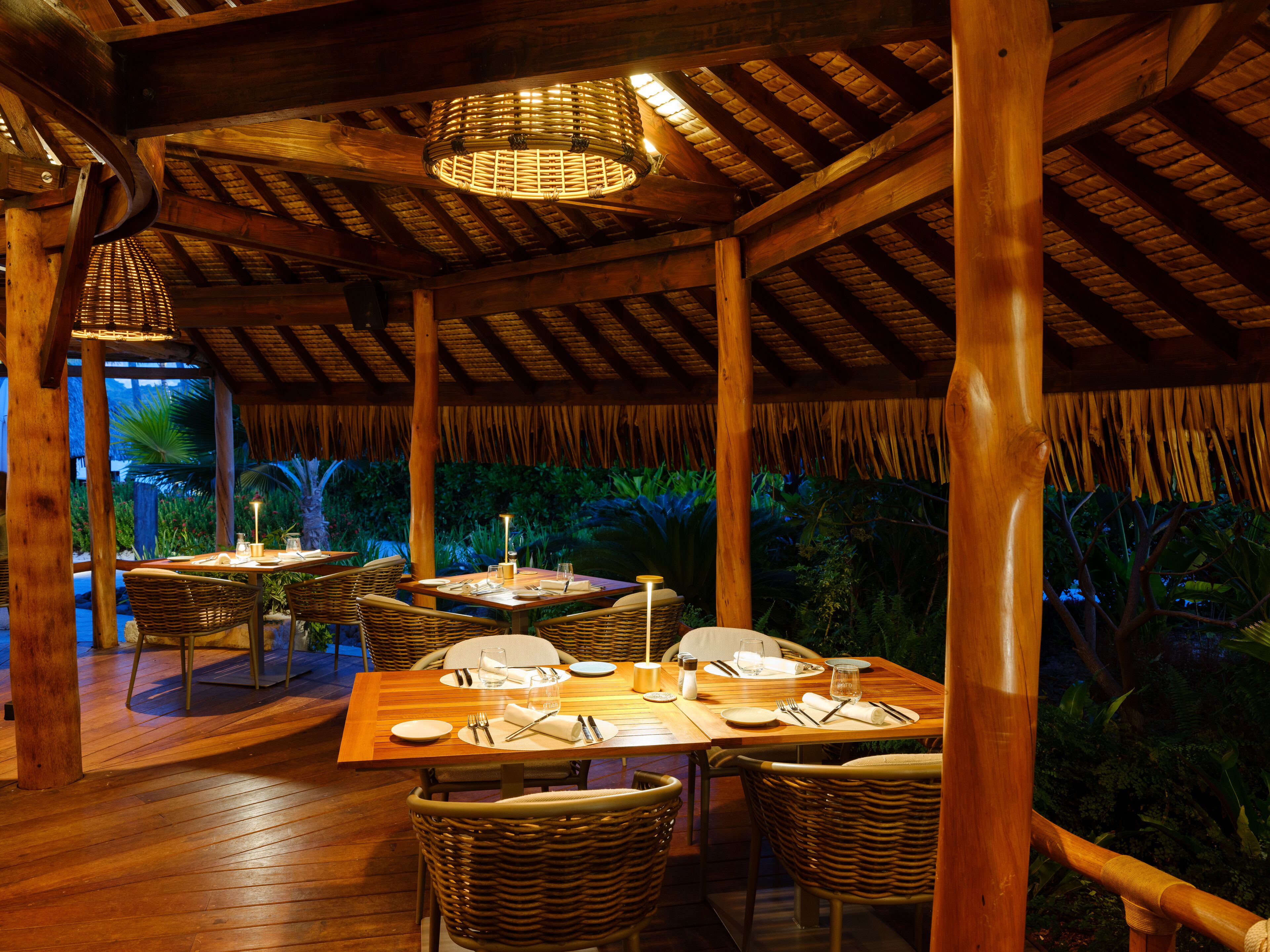 An outdoor dining area with wooden tables and wicker chairs, surrounded by lush greenery.