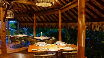 An outdoor dining area with wooden tables and wicker chairs, surrounded by lush greenery.